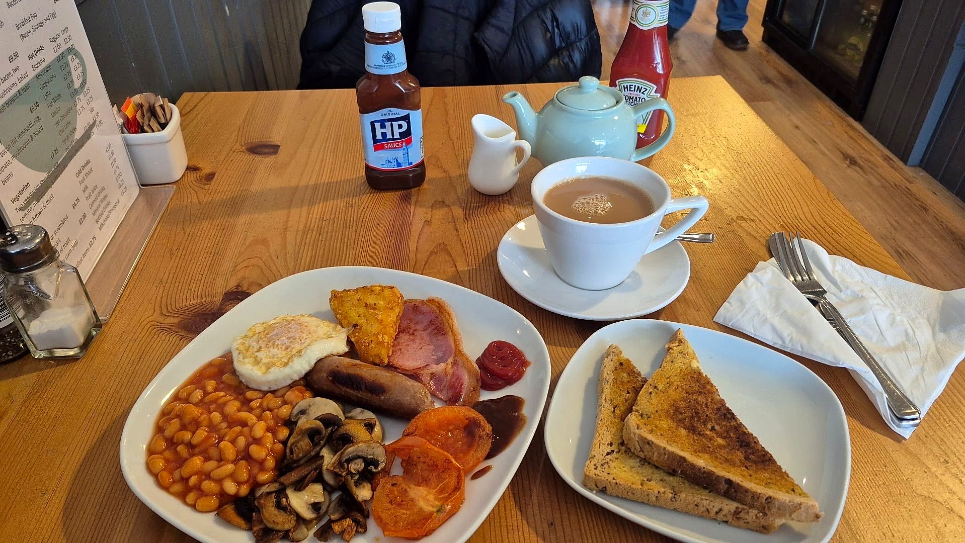 A cooked breakfast on a wooden table — egg, bacon, sausage, beans, mushrooms, tomato and hash brown — with toast, a pot of tea and a bottle of HP sauce