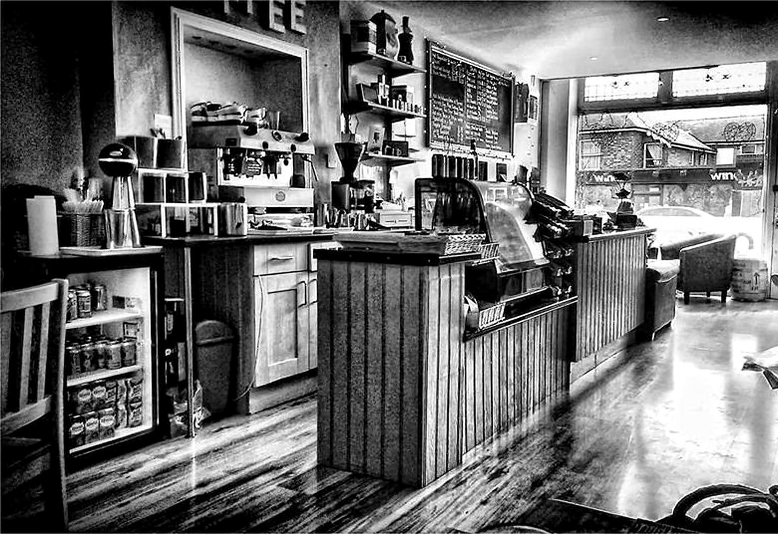 Black-and-white photo inside the café: a wood-panelled counter, coffee machine, shelves, a chalkboard menu and a polished wooden floor leading to the front window
