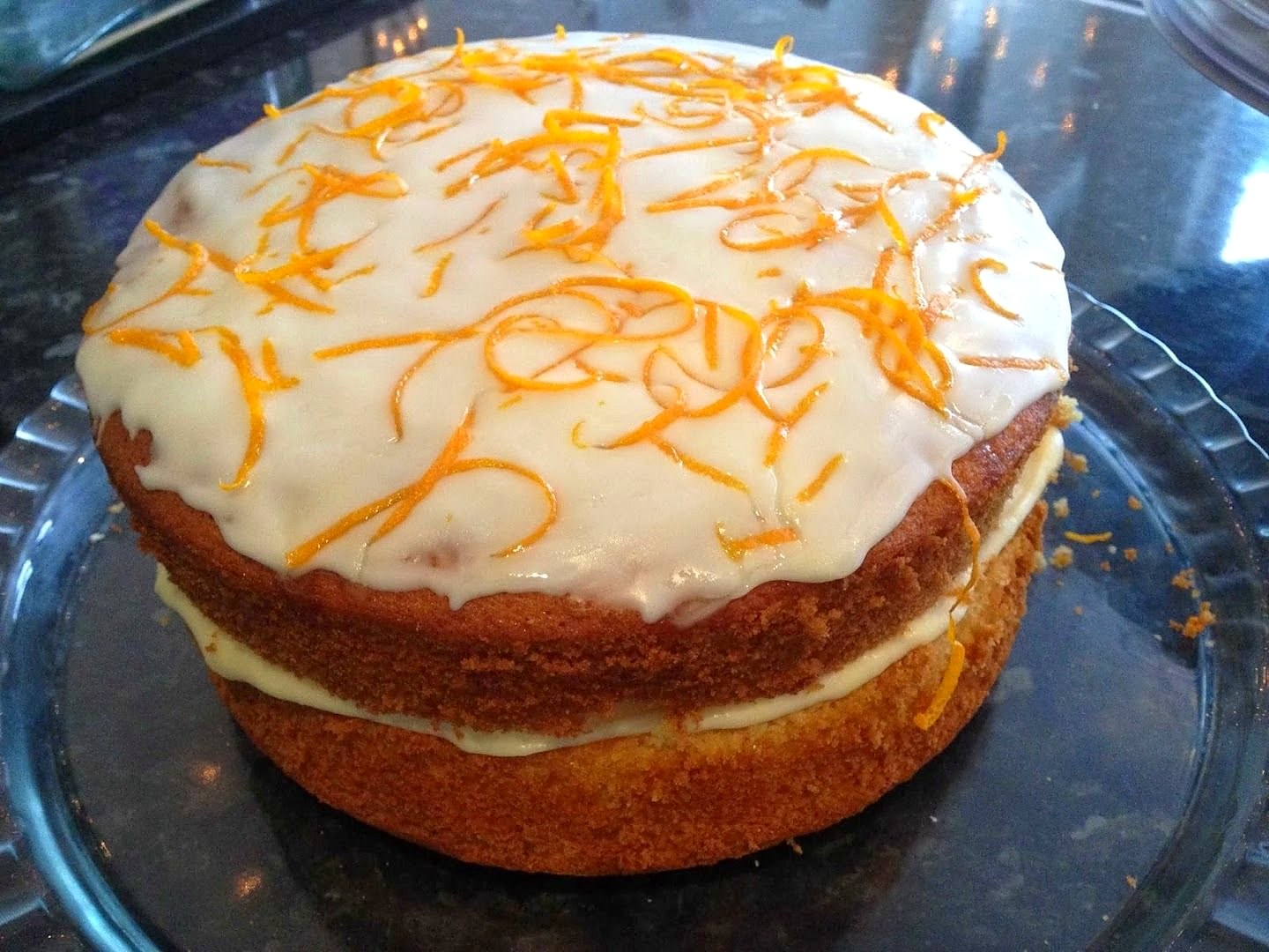 A whole two-layer sponge cake with white icing and a tangle of orange zest on top, on a glass cake stand